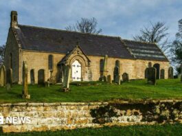 A small church with a white door. It sits in a graveyard, in a raised area with a wall in the foreground of the image.