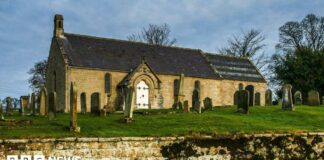 A small church with a white door. It sits in a graveyard, in a raised area with a wall in the foreground of the image.