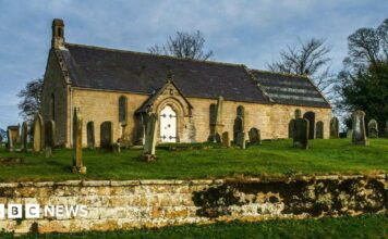 A small church with a white door. It sits in a graveyard, in a raised area with a wall in the foreground of the image.