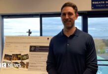 A man with short brown hair and a dark blue long-sleeved polo shirt stands in front of a sign promoting the redevelopment of Sixways Stadium.