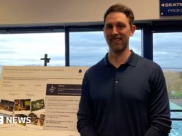 A man with short brown hair and a dark blue long-sleeved polo shirt stands in front of a sign promoting the redevelopment of Sixways Stadium.
