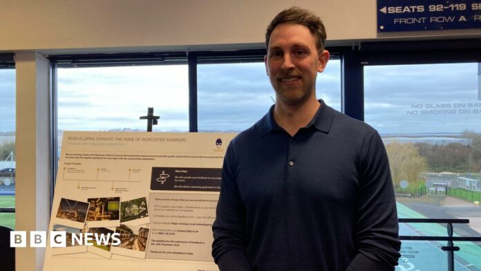 A man with short brown hair and a dark blue long-sleeved polo shirt stands in front of a sign promoting the redevelopment of Sixways Stadium.