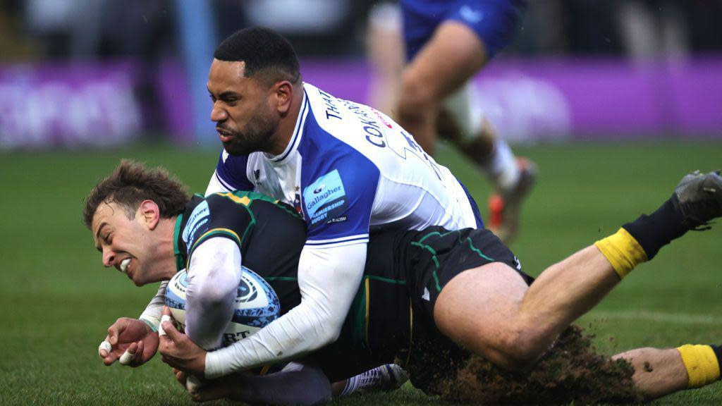 blue and white tackler tries to prevent player in green, black and yellow placing the rugby ball beyond the try line, legs in background