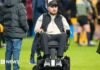 Former Gloucester Rugby player Ed Slater after the final whistle in The 745 Game at AMT Headingley Stadium, Leeds. He is wearing a black 4Ed hat, a black gilet, a beige fleece and black trousers. Ed is sat in a wheelchair on the pitch. Other people can be seen standing around him and nearby.