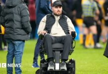 Former Gloucester Rugby player Ed Slater after the final whistle in The 745 Game at AMT Headingley Stadium, Leeds. He is wearing a black 4Ed hat, a black gilet, a beige fleece and black trousers. Ed is sat in a wheelchair on the pitch. Other people can be seen standing around him and nearby.