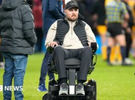 Former Gloucester Rugby player Ed Slater after the final whistle in The 745 Game at AMT Headingley Stadium, Leeds. He is wearing a black 4Ed hat, a black gilet, a beige fleece and black trousers. Ed is sat in a wheelchair on the pitch. Other people can be seen standing around him and nearby.