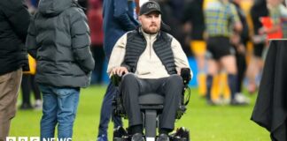 Former Gloucester Rugby player Ed Slater after the final whistle in The 745 Game at AMT Headingley Stadium, Leeds. He is wearing a black 4Ed hat, a black gilet, a beige fleece and black trousers. Ed is sat in a wheelchair on the pitch. Other people can be seen standing around him and nearby.