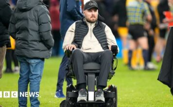 Former Gloucester Rugby player Ed Slater after the final whistle in The 745 Game at AMT Headingley Stadium, Leeds. He is wearing a black 4Ed hat, a black gilet, a beige fleece and black trousers. Ed is sat in a wheelchair on the pitch. Other people can be seen standing around him and nearby.