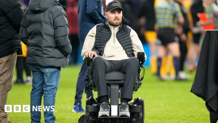 Former Gloucester Rugby player Ed Slater after the final whistle in The 745 Game at AMT Headingley Stadium, Leeds. He is wearing a black 4Ed hat, a black gilet, a beige fleece and black trousers. Ed is sat in a wheelchair on the pitch. Other people can be seen standing around him and nearby.