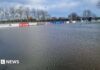 Wide shot of rugby pitches completely submerged, with goal posts and advertising boards the only items visible. Trees are in the background. 