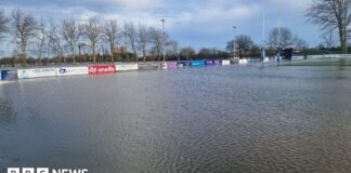 Wide shot of rugby pitches completely submerged, with goal posts and advertising boards the only items visible. Trees are in the background. 