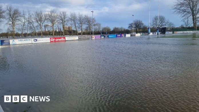 Wide shot of rugby pitches completely submerged, with goal posts and advertising boards the only items visible. Trees are in the background. 