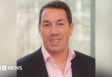 A head and shoulders shot of Tom Voyce. He is wearing a dark suit jacket and a pink shirt. He's clean shaven, in his forties, with dark hair and is smiling 