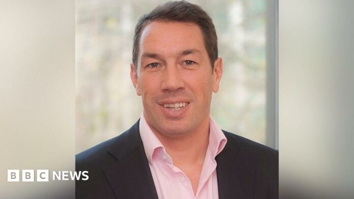 A head and shoulders shot of Tom Voyce. He is wearing a dark suit jacket and a pink shirt. He's clean shaven, in his forties, with dark hair and is smiling 