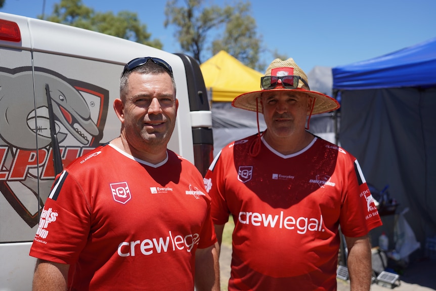 Two men in matching rugby shirts near a van on a sunny day.