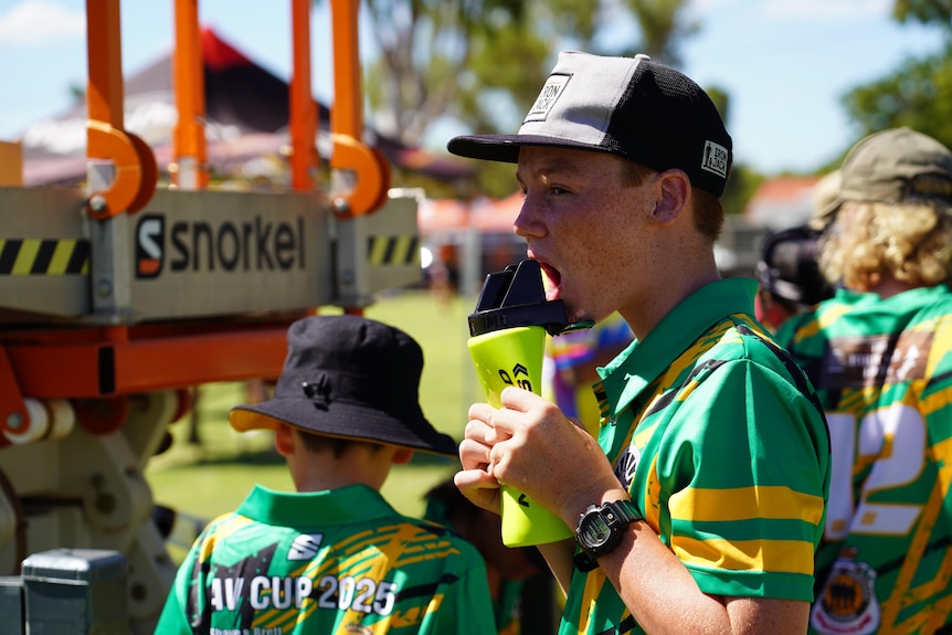 A boy in a rugby league uniform drinks from a bottle at a sports ground on a sunny day.