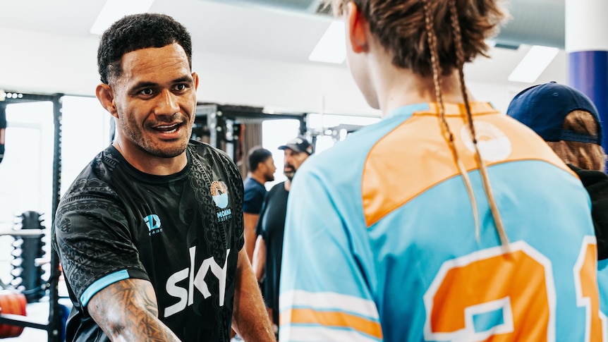 A man with a tribal tattoo sleeve wearing a black rugby jersey shakes the hand of a european boy with two plaits in his hair