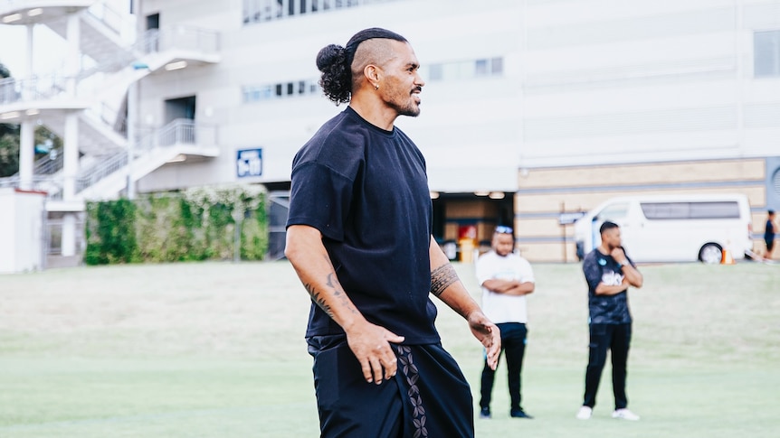 Side profile of a man wearing black shirt and black ie lavalava and black shoes with arms slightly raised and hair in a bun