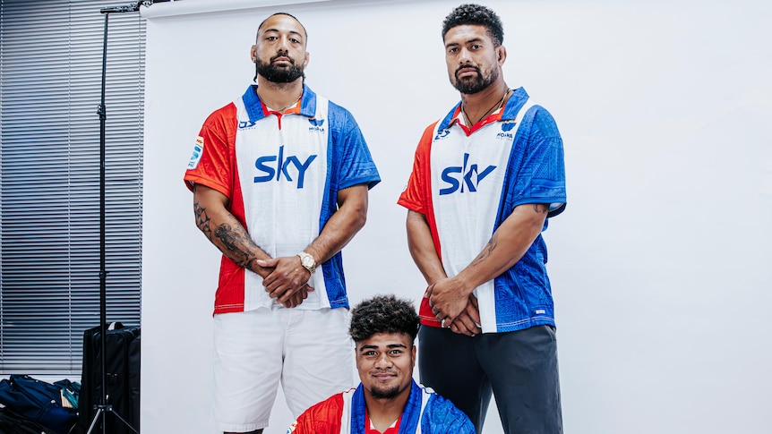 3 Pacific island men pose in a studio wearing red, white & blue large stripe rugby jerseys.  One man sits cross legged on floor